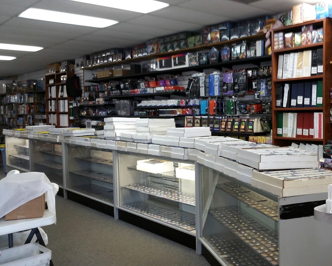 The length of a card shop counter, glass cases of single cards below and boxed product stacked to the ceiling behind.